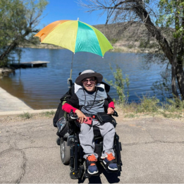 A young man in a wheelchair thoughtfully enjoys the lake with an attached umbrella.