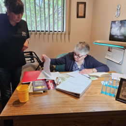An older woman writing letters as a staff person supervises